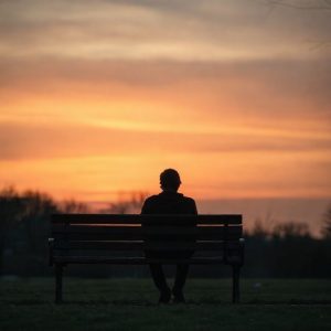 A person sitting alone on a park bench during sunset, looking contemplative. The sky has warm, melancholic colors. The mood is calm and reflective.