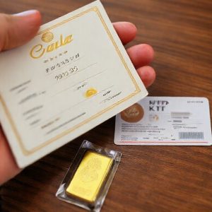 Close-up of a hand holding a Galeri 24 gold certificate, showing details like weight and purity, next to a KTP card on a wooden table. A small gold bar is partially visible.